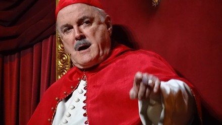 A close-up of actor John Cleese dressed in Roman papal attire, and pointing at the camera while mid-speech.