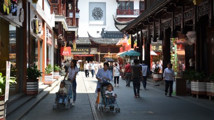 People walking through a shaded road in Shanghai pushing prams, with traditional Chinese architecture on either side.