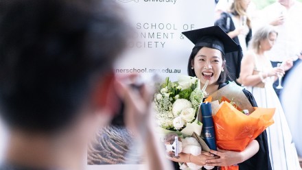 A Fenner graduate stands in front of a banner and smiles while people in the foreground take photos.
