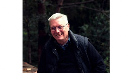 Dr Ian Fry is wearing glasses, a dark zippered vest and black top underneath, with a dark background. His hair is short and grey and he is smiling.