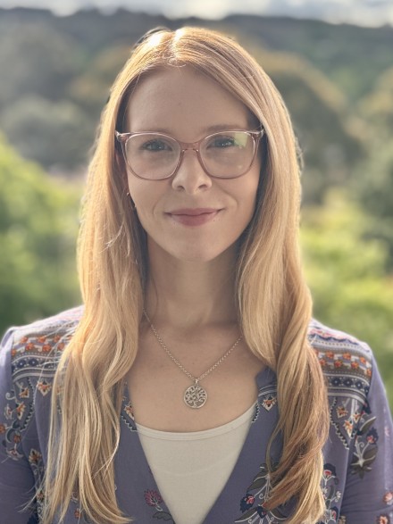A picture of Dr Elle Bowd. She wears framed glasses, and has long gold-blonde hair. Behind her head and shoulders is a blurred bushland landscape.