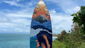 A surfboard covered in an ocean-scape and filled with Pasifika flowers stands up, pointing to a blue sky.