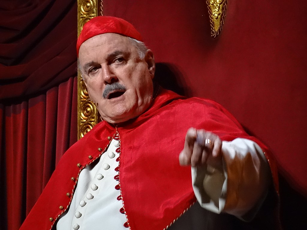 A close-up of actor John Cleese dressed in Roman papal attire, and pointing at the camera while mid-speech.