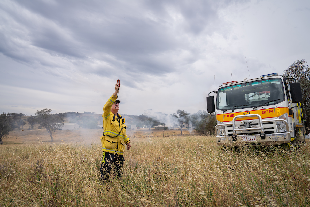 A fire fighter holds up a remote control to the sky with a fire truck behind him.