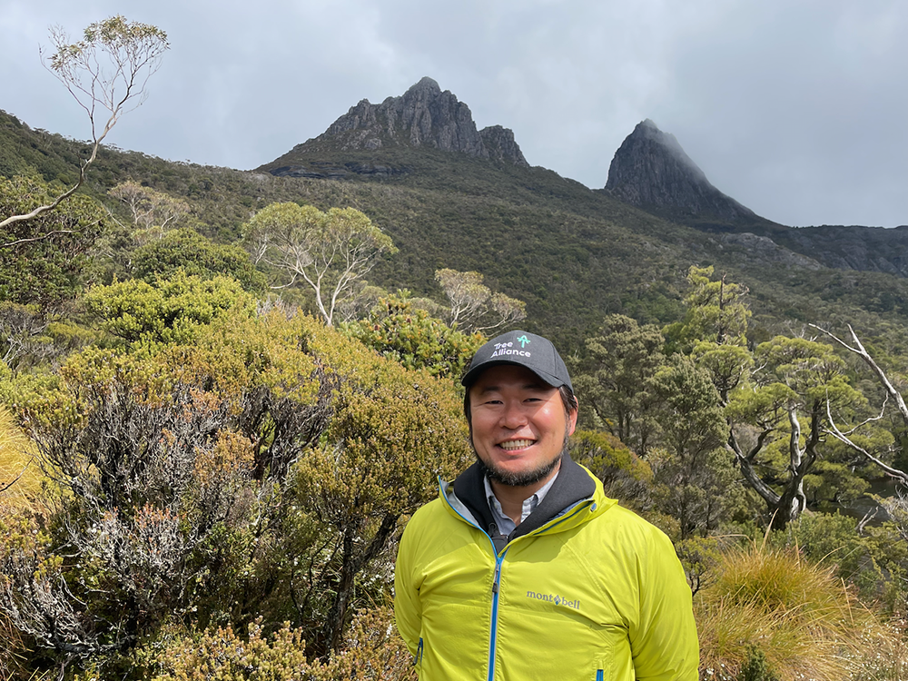 Kaz stands in front of bushland, he wears a hat, a yellow jacket and is smiling at the camera.
