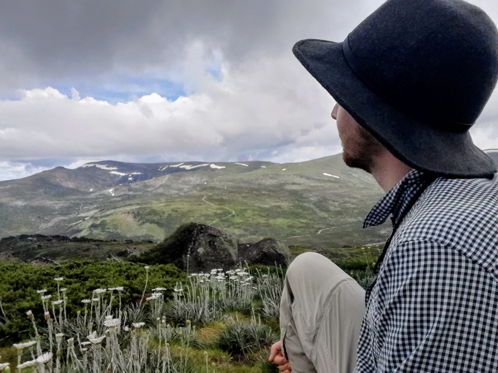 Josh sits on a grassy alpine slope, facing away from the camera and looking out over a mountainous landscape with patches of snow and cloud-covered skies. They wear a wide-brimmed dark hat, a checkered long-sleeve shirt, and light-coloured pants. In the foreground, native alpine plants with tall white flower stalks are visible, adding texture to the natural scene