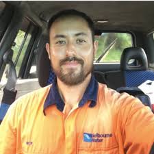 Portrait photo of Nicholas Metherall wearing an orange high-visibility work shirt with a 'Melbourne Water' logo, seated inside a vehicle, smiling at the camera.