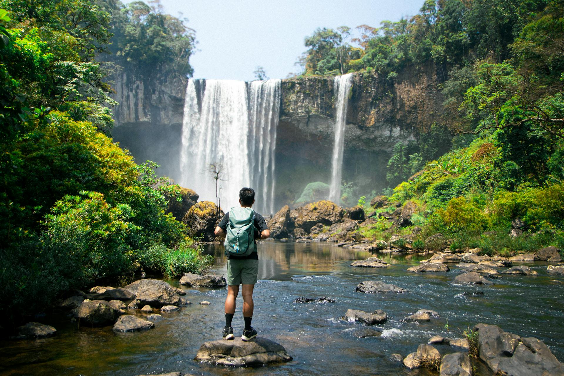 A person wearing a green backpack stands on a rock in the middle of a shallow river, facing a large waterfall that cascades from a tall cliff surrounded by lush, tropical forest. The individual appears to be hiking or exploring, dressed in outdoor gear including a short-sleeved shirt, shorts, and socks with sneakers. Bright sunlight filters through the trees, highlighting the greenery and the mist from the waterfall.