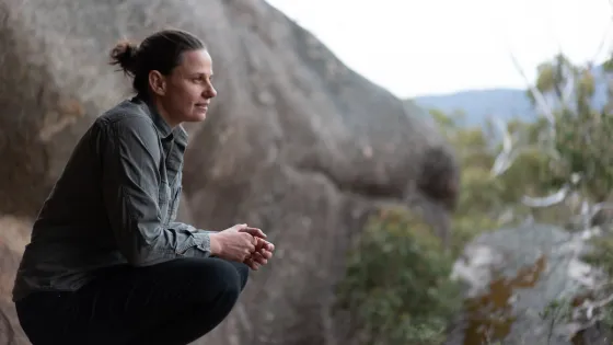 A person sitting and looking contemplatively in a natural outdoor setting with rocks and foliage.