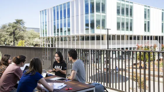 A group of students sit around a table outside a modern university building, engaged in discussion.
