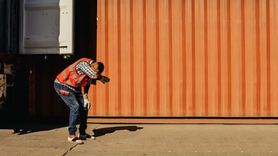 A man stands in workwear in front of an orange wall outside, on concrete. He has his head in his hands and his hunched over his knees.