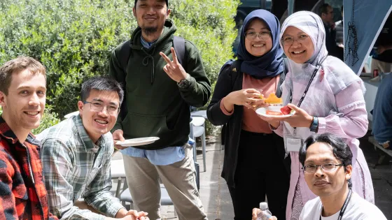 A group of six people smiling and interacting during what appears to be an outdoor event, some holding plates with food.
