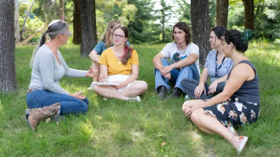 A group of five adults sitting in a grassy area under trees, engaged in a discussion.