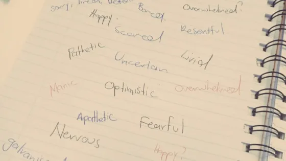 A notepad with handwritten words describing various emotions such as "resentful," "curious," "overwhelmed," and "furious," alongside pens and other items on a desk.