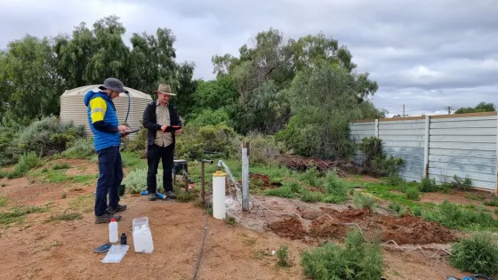 Two men examine equipment at a rural outdoor site with sparse vegetation and a water tank in the background.