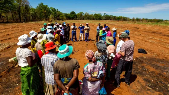 A group of men and women, many wearing hats, gather for a meeting in a large open field under a clear blue sky.
