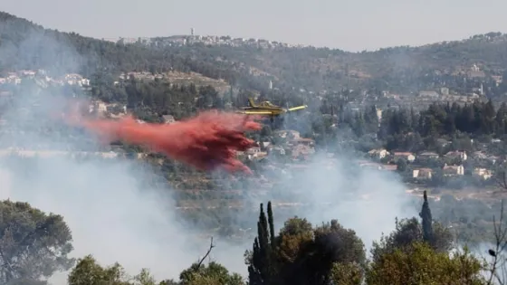 A yellow firefighting aircraft drops red retardant on a forested area to combat a wildfire.