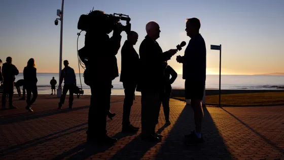 Silhouetted figures conducting a television interview on a boardwalk at sunset, with the ocean in the background.