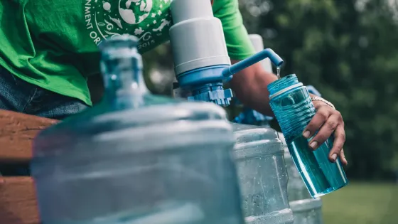 A person in a green environmental organization t-shirt refilling a reusable water bottle from a larger water container outdoors.