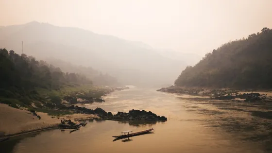A serene river scene with a boat on calm waters, surrounded by forested mountains and hazed by golden light.