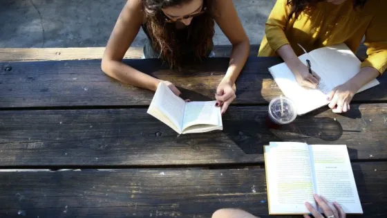 Two women studying and taking notes at a wooden table outdoors.