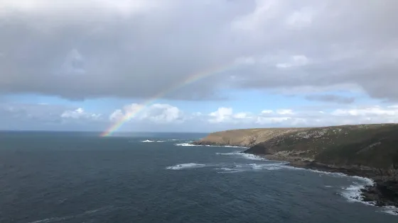 A panoramic coastal scene showing a rugged shoreline and open ocean beneath a mostly cloudy sky. A faint rainbow arcs from the water on the left toward a grassy headland on the right. Waves crash gently along the rocky edge of the coast, and scattered patches of blue sky peek through the clouds. The overall mood is calm and atmospheric.