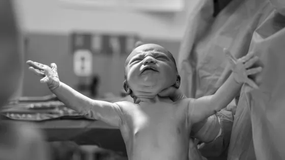 A newborn baby stretches its arms out while being held by a medical professional in a hospital setting.