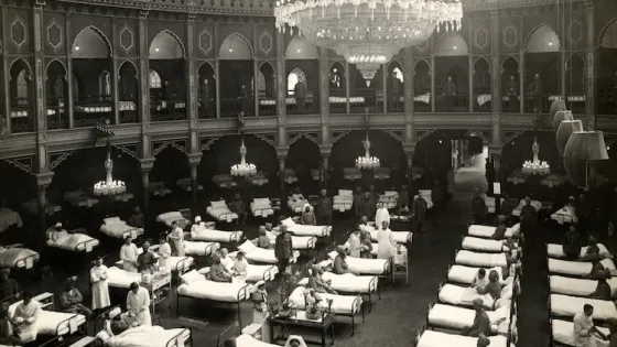 A historical photo of a large ornate hall transformed into a hospital ward with rows of beds and patients, illuminated by a grand chandelier.
