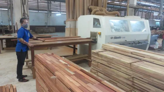A worker in a woodworking factory handles lumber next to large industrial machinery.