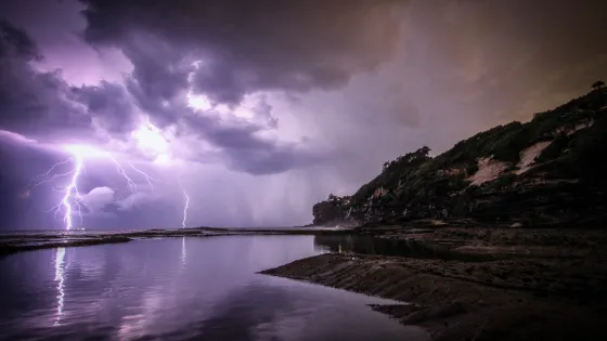 A dramatic nighttime scene on a coast with multiple lightning bolts striking near the water and dark, stormy clouds overhead.