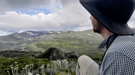 Josh sits on a grassy alpine slope, facing away from the camera and looking out over a mountainous landscape with patches of snow and cloud-covered skies. They wear a wide-brimmed dark hat, a checkered long-sleeve shirt, and light-coloured pants. In the foreground, native alpine plants with tall white flower stalks are visible, adding texture to the natural scene