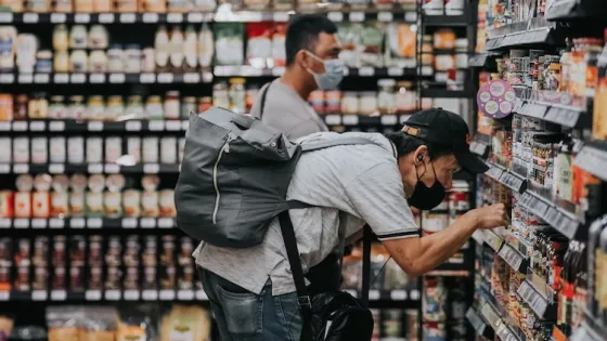 A man wearing a mask and a backpack is browsing products in a grocery store aisle.