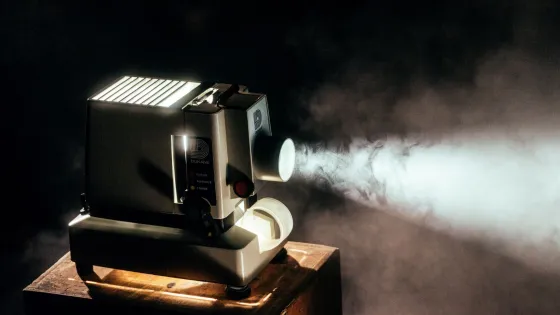 An old projector emitting light and smoke while sitting on a wooden surface in a dimly lit room.