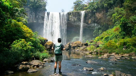 A person wearing a green backpack stands on a rock in the middle of a shallow river, facing a large waterfall that cascades from a tall cliff surrounded by lush, tropical forest. The individual appears to be hiking or exploring, dressed in outdoor gear including a short-sleeved shirt, shorts, and socks with sneakers. Bright sunlight filters through the trees, highlighting the greenery and the mist from the waterfall.