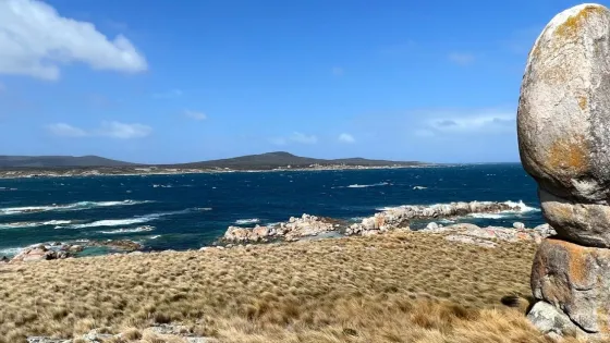 A coastal landscape featuring a strikingly tall and narrow balanced granite boulder standing upright on grassy terrain in the foreground. The scene overlooks a windswept ocean with white-capped waves and rocky outcrops. In the distance, rolling hills stretch across the horizon beneath a blue sky dotted with clouds. The location has a wild, remote feel.