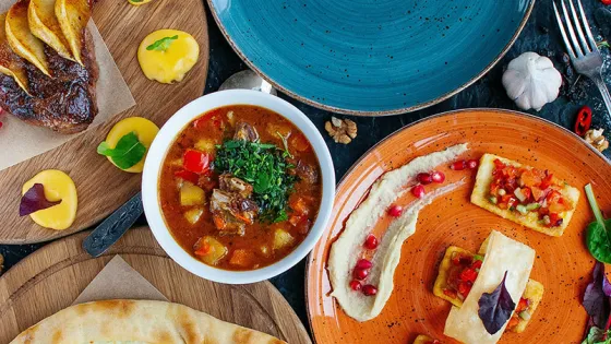 An overhead shot of a vibrant and assorted array of dishes including soup, steak with sauces, and a variety of appetizers on a textured dark background.