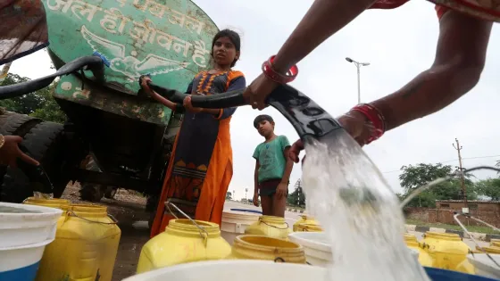 Children fill water containers from a large water tanker in a rural setting.