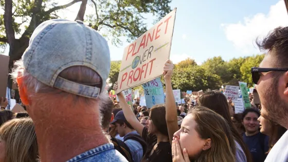 A crowd of people at a protest, with one person holding a sign reading "Planet Over Profit."
