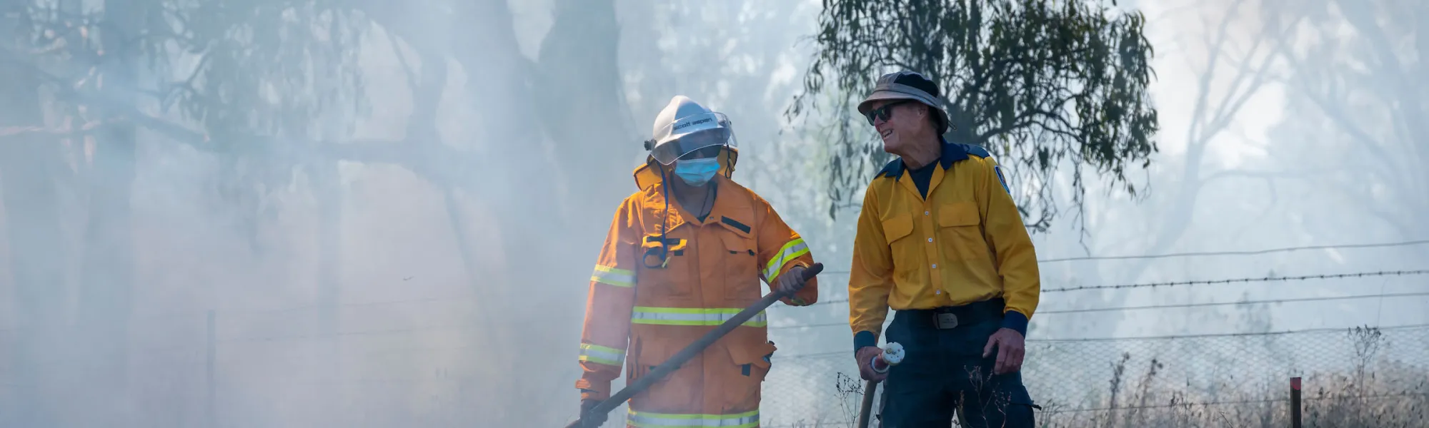 Two people in fire fighting attire face each other and smile while undertaking cultural burning in a woodland.