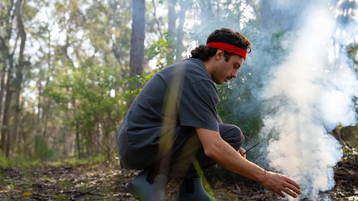 Adrian Webster crouches over a small fire on a forest floor, with plumes of smoke floating above it.