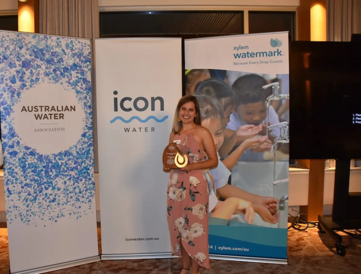 In a warmly-lit (and slightly dim) official-looking room, a young woman stands holding a drip-shaped award in gold. She is standing in front of three banners that all read 'ICON'. She is smiling, with blonde hair and a pale pink tulip-petal long dress.