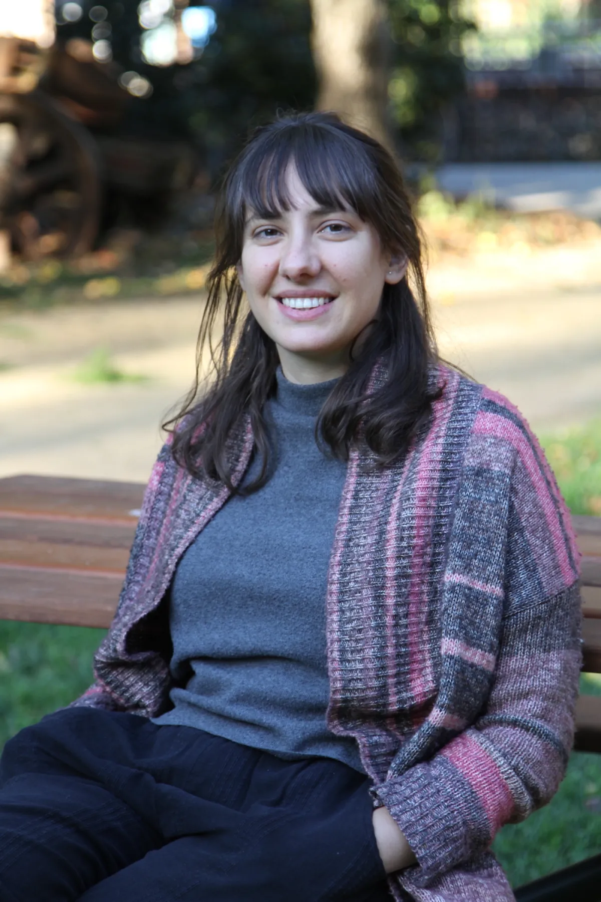 PhD graduate Dr Connie Leon sits on a wooden bench in a grey skivvy and pink and grey long cardigan. She has a blunt fringe, and a gentle smile on her face.