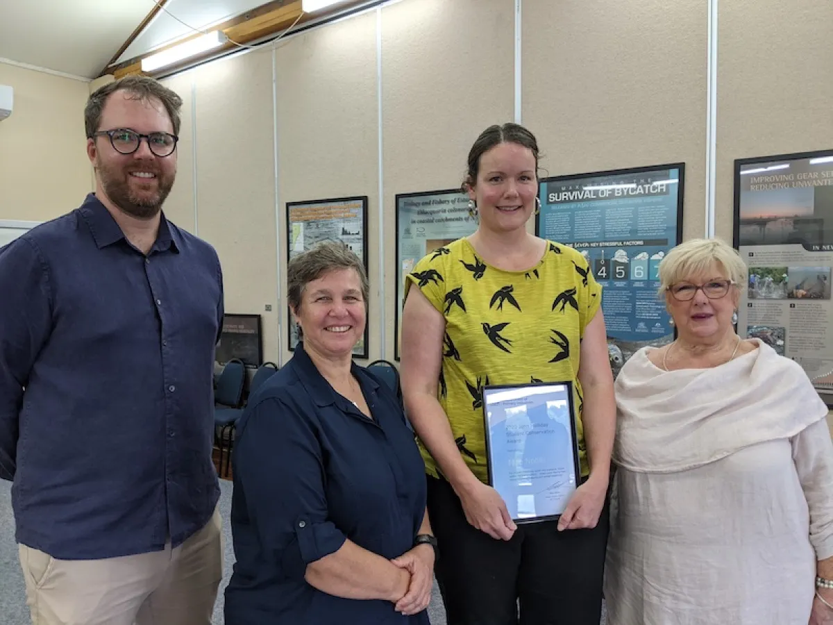 Four people stand together in a room. The second from the right is a young woman with a yellow t-shirt on, holding a framed award.