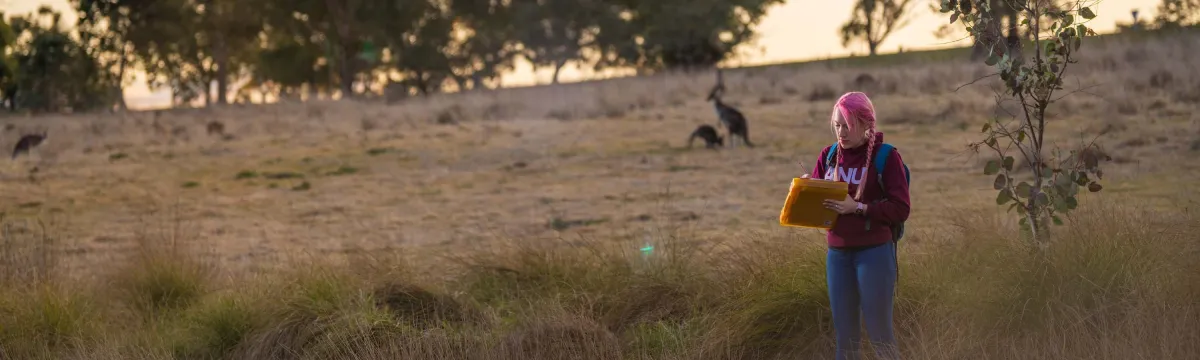 A student takes notes at the edge of a dam with eucalyptus trees and kangaroos in the distance.