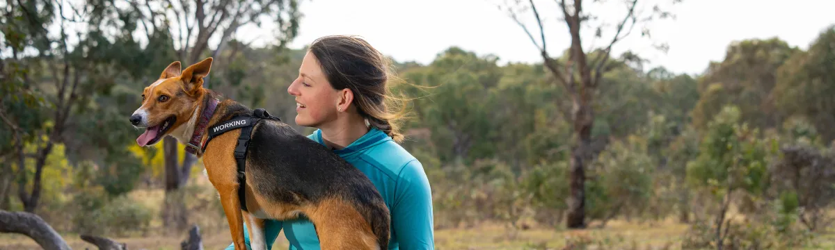 A Fenner researcher, Shoshana Rapley, kneels with her dog on her leg and looks at her, with woodland in the background.