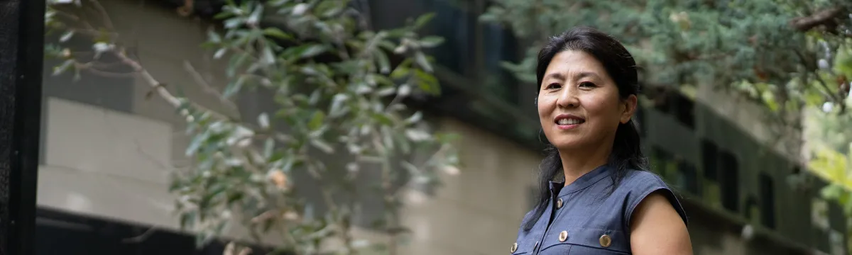 Researcher, Xuemei Bai, stands in front of the Fenner building with a grass tree beside her.