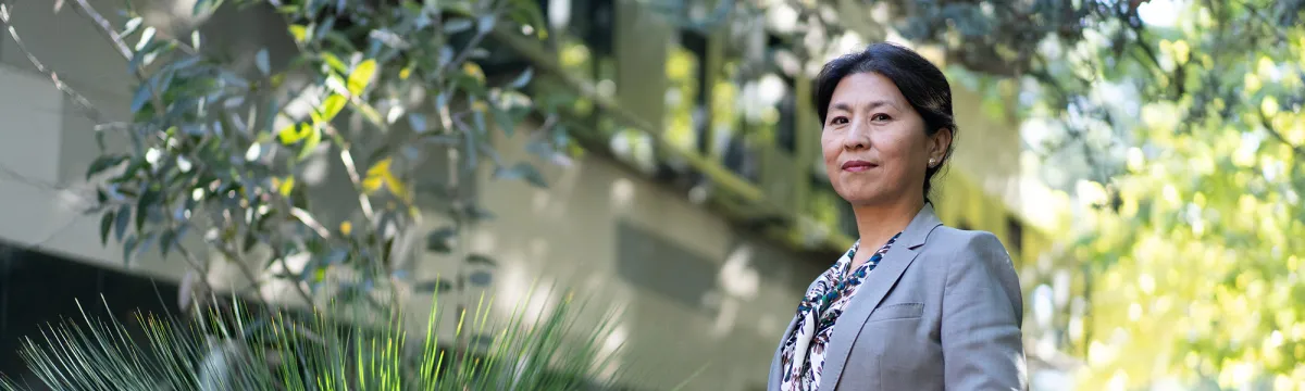 Researcher, Xuemei Bai, stands in front of the Fenner building with a grass tree beside her.