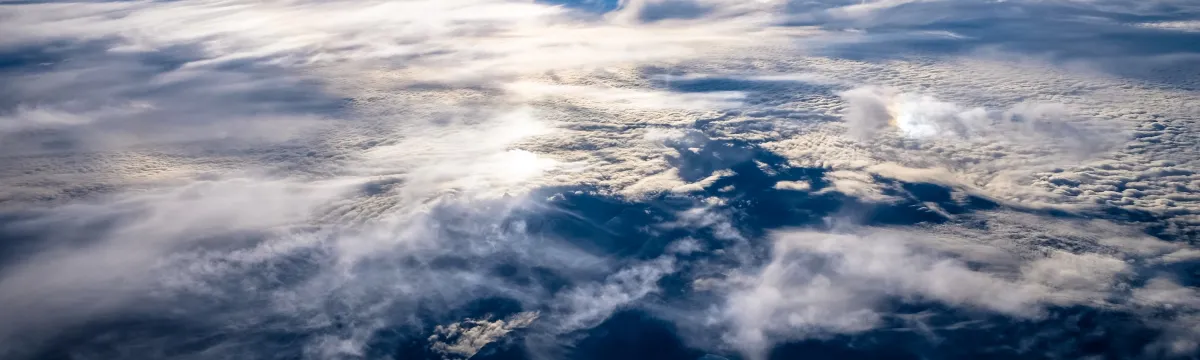Aerial view above a dense layer of clouds, with soft, wispy textures across the frame. Sunlight filters through the upper atmosphere, casting bright reflections on the cloud tops. The sky above is deep blue, gradually lightening toward the horizon. Below the clouds, dark mountainous terrain is faintly visible through gaps in the cloud cover, creating a sense of altitude and vastness.