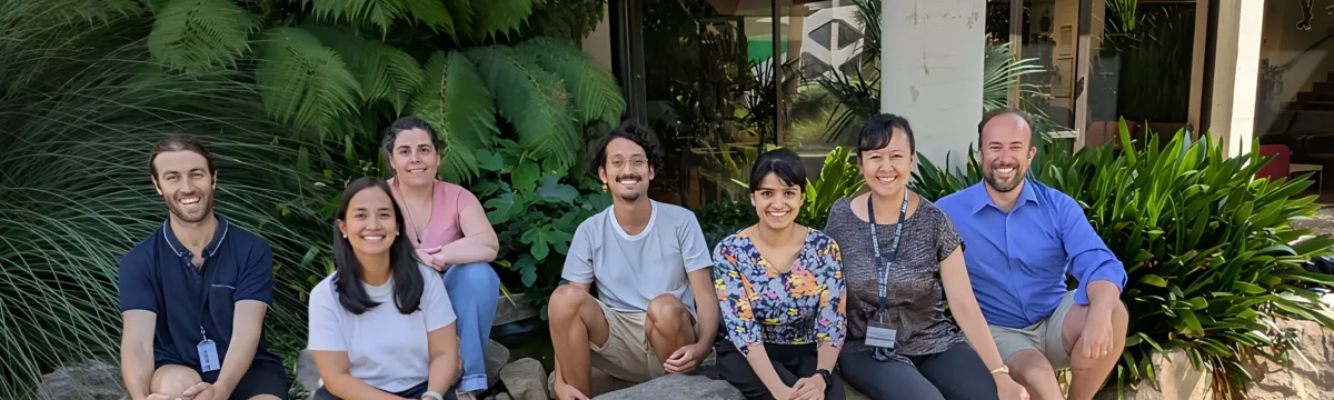 A group of researchers sit among large rocks and ferns outside a building.
