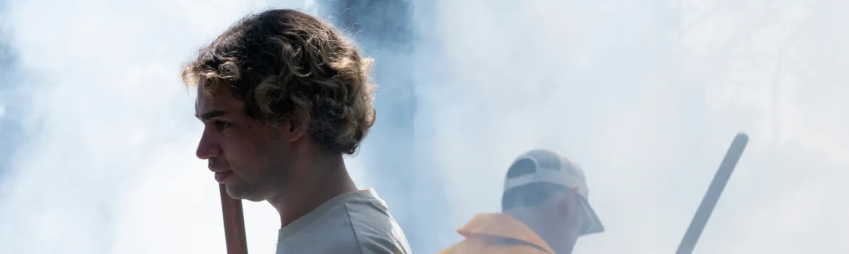 A man walks through a smoky woodlands with someone standing in the background in firefighting gear.